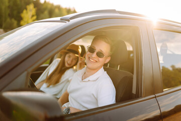 Young couple in a new car is relaxing in the sunset. Man has fun with a girl in a car at the wheel....