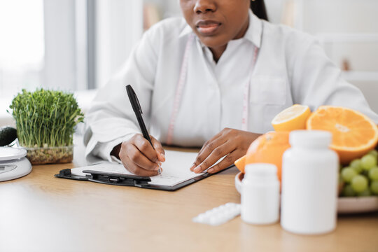 Close up view of multiracial female in white coat filling in paper form on clipboard while staying indoors. Food expert devising individual meal plan due to personal preferences of patient at work.