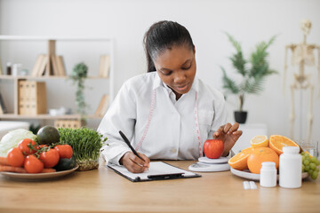 Focused multiracial woman in lab coat taking notes while assessing apple via dietary scale in...