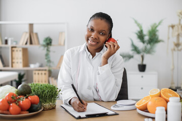 Portrait of smiling female posing with apple close to cheek while doing paperwork in doctor's...