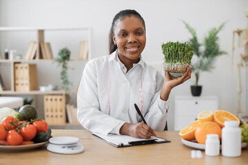 Portrait of joyful female in lab coat posing with green sprouts of herbs in hand while using writing surface at desk. Multicultural nutritionist creating well-balanced diet containing microgreens.