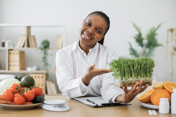 Smiling multicultural woman gesturing at leafy seedlings in container while staying at work in...