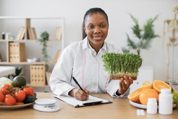 Positive female nutritionist with young sprouts in hand creating meal diet at writing desk in...