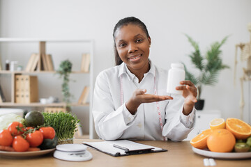 Cheerful multiracial woman offering pills container at camera during office hours in modern clinic....