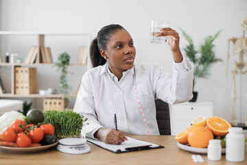Focused african american woman in white coat checking fluid clearness in glass at doctor's workplace. Effective nutrition specialist advising about drinking water for boosting metabolism at diet.
