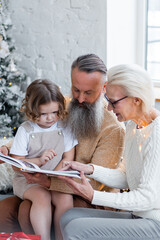 Senior grandparents, attractive grandmother and handsome grandfather reading a fairy tale book to cute little granddaughter near decorated Christmas tree. Two generations, family values, happy people