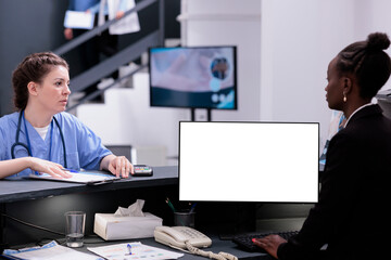 African american receptionist using pc with white isolated screen at reception counter desk in hospital. Diverse medical staff working with mockup template during checkup visit consultation