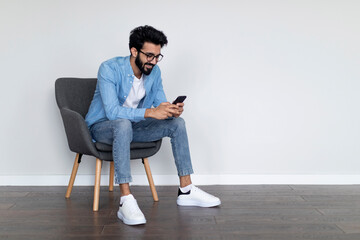 Cheerful Young Indian Man Using Cellphone While Sitting In Armchair At Home