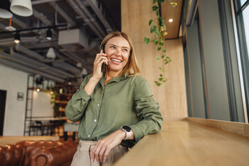 Smiling woman manager is talking phone with client while standing on modern coworking background