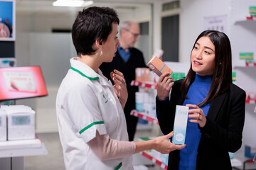Pharmacist recommending young asian woman sunblock cream in drugstore. Customer holding skin care products packages, choosing between two sunscreen lotions and consulting with pharmacy assistant