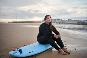 Fototapeta premium Dreamy body positive surfer woman looking away, while sitting with surf board