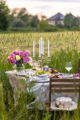Elegant fancy wedding table decor with bright ranunculi flowers in a vase outdoors, chandelier, field or meadow. Summer, sunset, golden hour. Romantic dinner for a couple in restaurant outside