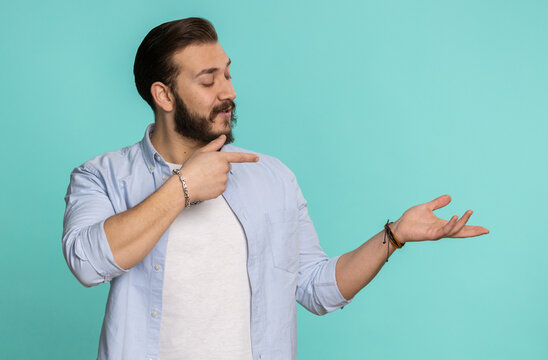 Young lebanese man showing thumbs up and pointing empty place, advertising area for commercial text, copy space for goods promotion. Bearded arabian guy isolated on blue studio background, indoors