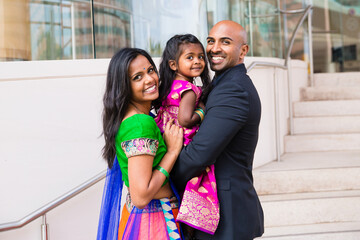 beautiful indian family with daughter girl hugging and smiling with a bindi and traditional sari dress in front of a building with staircase