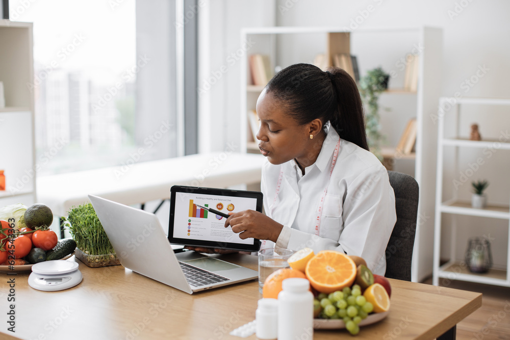 Wall mural Focused multicultural expert with meal plan on tablet conducting web meeting using portable laptop at work. Skilful female nutritionist explaining details of slimming program for client online.