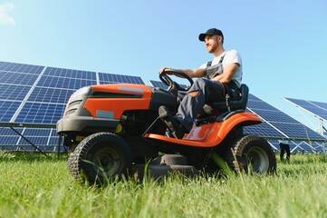 Fototapeta na wymiar A man working at solar power station. A worker on a garden tractor mows grass on a solar panel farm.