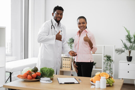Multiracial specialist taking measurements of lady's waist at dietitian's office in clinic. Attentive male professional evaluating client's health by estimating body composition, showing thumbs up