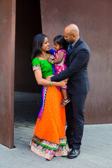 Fototapeta na wymiar beautiful indian family with daughter girl hugging and smiling with a bindi and traditional sari dress in front of a rust colored building