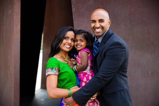 beautiful indian family with daughter girl hugging and smiling with a bindi and traditional sari dress in front of a rust colored building