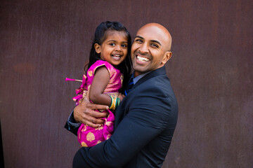 beautiful indian family dad father with daughter girl hugging and smiling with a bindi and traditional sari dress in front of a rust architectural building
