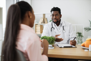 Confident man with stethoscope writing on paper clipboard during conversation with multiracial woman in office. Serious nutrition expert making notes of patient's medical history for proper meal plan.