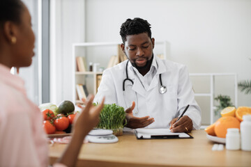 Confident man with stethoscope writing on paper clipboard during conversation with multiracial woman in office. Serious nutrition expert making notes of patient's medical history for proper meal plan.