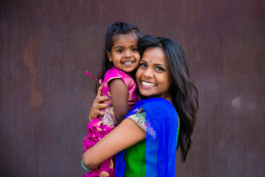 beautiful indian family mom mother with daughter girl hugging and smiling with a bindi and traditional sari dress in front of a rust architectural building