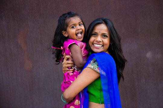 beautiful indian family mom mother with daughter girl hugging and smiling with a bindi and traditional sari dress in front of a rust architectural building