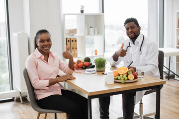 Full length portrait of multicultural people posing at office desk in doctor's workplace of medical center. Attractive woman in casual wear visiting professional dietitian, showing thumbs up