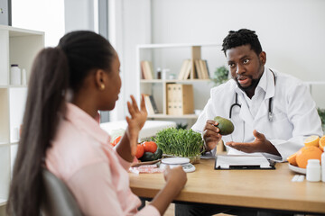 Focused african american man in white coat holding avocado while talking with female client in...