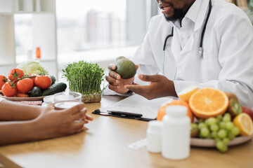 Close up view of bearded man holding green fruit while chatting with multiethnic person in office interior. Male nutritionist describing benefits of slimming foods during appointment in modern clinic.