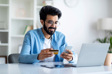 E-Commerce Concept. Smiling Indian Man Holding Credit Card And Using Cellphone