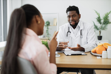 Attractive bearded man in doctor's coat gesturing at glass with fluid while meeting with multiethnic patient. Smiling nutritionist recommending clean water as help in reducing calorie intake on diet.