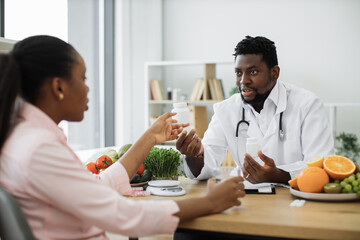 Handsome african american man in white coat giving pills bottle to charming woman in consulting room of hospital. Male nutrition professional improving overall health with dietary supplements.