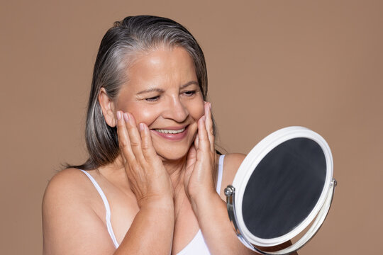 Cheerful elderly caucasian lady with gray hair touches face with hands, look in mirror, enjoy anti-aging spa treatment
