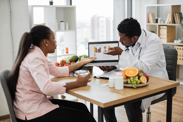 African american doctor and patient having appointment in hospital cabinet and discussing woman's treatment. Long haired lady holding glass of water while male specialist pointing at graphs on laptop.