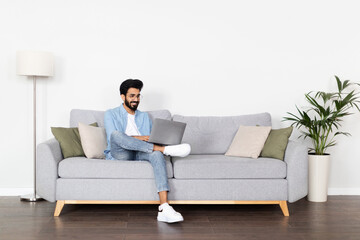 Positive arab guy using laptop, sitting on couch at home