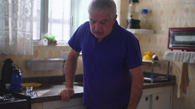 Lonely unshaven senior man standing at home leaning on sink by kitchen with depressed expression. worn-down person struggling with old age