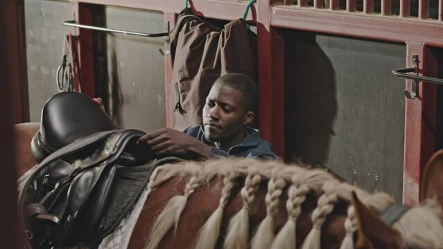 High angle shot of young black man putting saddle on horse with braided mane in stable while preparing for ride