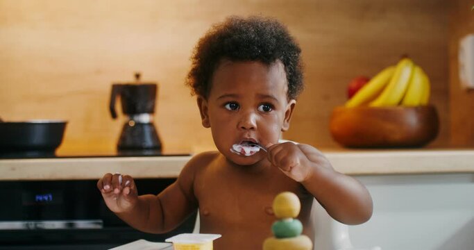 A baby of African-American appearance is eating yogurt with a spoon sitting in a chair, drops of yogurt dripping on her naked tummy
