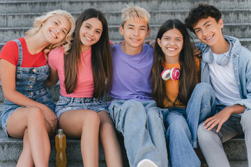Group of smiling friends, stylish multiracial teenagers sitting on stairs looking at camera . Happy boys and girls communication, relaxing on the street. Friendship, positive lifestyle, summer