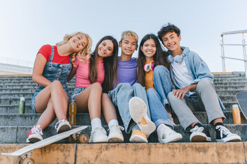 Portrait of smiling multiracial teenagers sitting on stairs looking at camera on urban street. Happy friends hugging together outdoors Friendship, diversity concept 