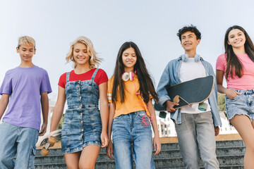 Obraz premium Group of smiling multiracial teenage friends wearing colorful t shirts talking, communication, holding skateboards walking on the street. Happy stylish boys and girls outdoors. Friendship concept