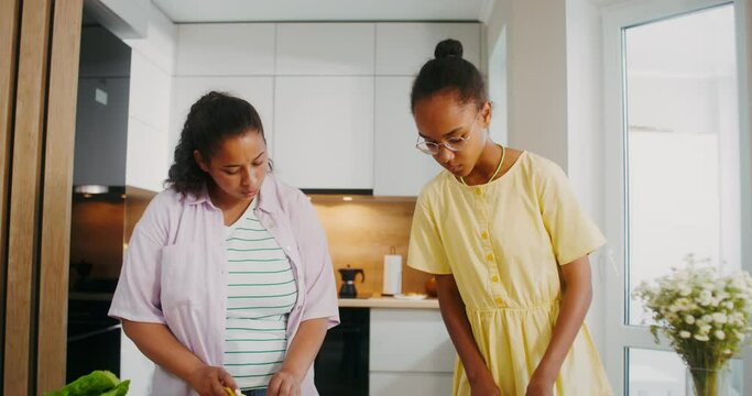 The baby is watching cartoons on a tablet while sitting in a child's chair while her mother and older sister prepare a salad standing at the table next to her
