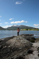 A girl stands on a stone on the seashore and looks at the mountains on a sunny day, Northern Norway