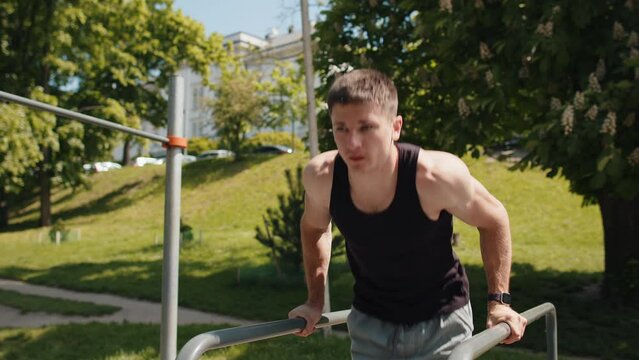 Athletic caucasian man in sportswear practicing dips on parallel bars, training triceps. Young guy in t-shirt on playground. Sports health fitness routine, workout. Strength motivation. Outdoor gym