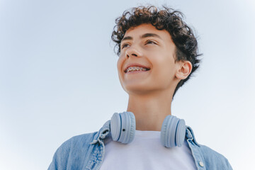 Portrait of smiling curly haired teenager with braces wearing headphones looking away standing on...