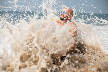 Smiling vacationist in sunglasses covered by wave sitting on sandy seaside. Splash of water scattered on foreground.
