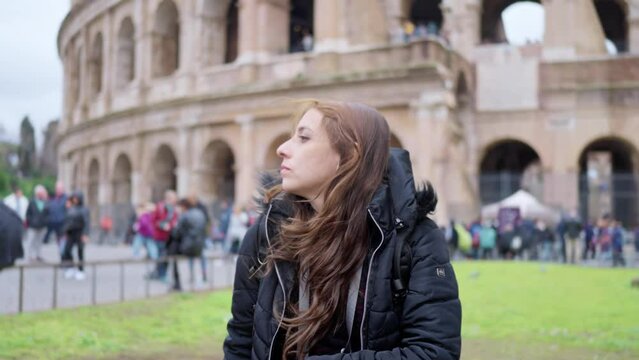 Positive young Latin woman tourist in warm outfit looking away against Colosseum during trip through Rome, Italy