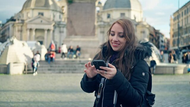 Smiling young Latin woman tourist with warm clothes using smartphone and professional photo camera while standing in Piazza del Popolo in Rome, Italy
